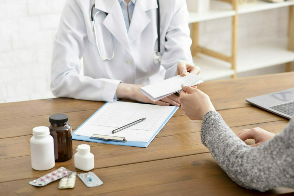 Prescription for medication and examination by doctor. Adult woman therapist gives paper to patient at workplace with tablet and jars of pills in office interior, cropped, close up, copy space